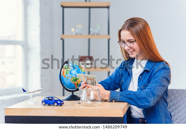 Young beautiful girl takes money from\
glass jar on the wooden table with globe, toy car and model of air\
plane on the white room background. Close up\
shot.