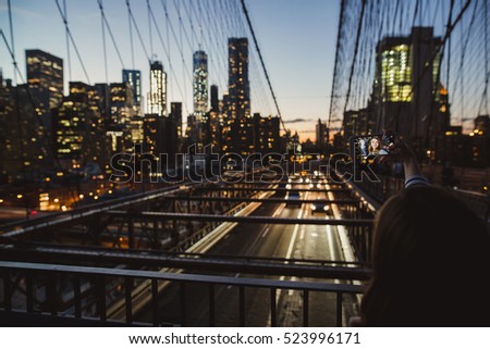 Young beautiful girl taking selfie with mobile phone on at Brooklyn Bridge at night. Amazing view of New York on the background.