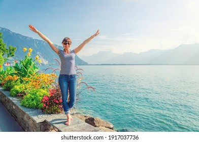 Joven y hermosa viajera con camisa a rayas y jeans posando, levantando las manos y sonriendo en el terraplén del lago Leman Ginebra en la ciudad de Montreux de la Riviera Suiza, montaña Alpes de fondo, Suiza