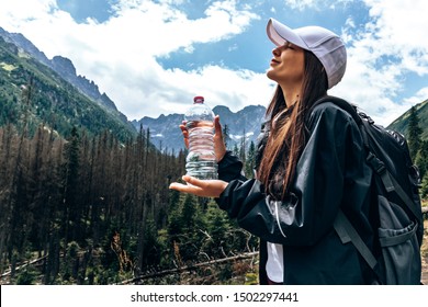 Young beautiful girl traveler in white hat and black jacket and backpack, standing with closed eyes and holding a bottle of water, amazing mountains on the background, summer time