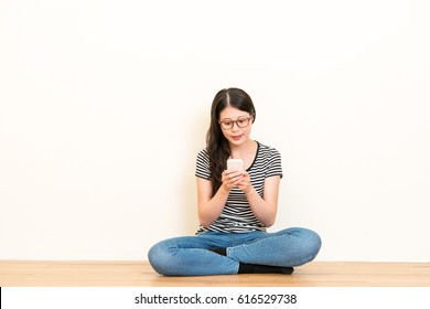 Young Beautiful Girl Using Cell Phone Facetime Or Typing Chatting With Family Sitting On Wooden Floor Over Blank Copyspace White Wall Background.