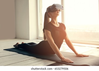 A Young Beautiful Girl In Virtual Reality Glasses Makes Yoga And Aerobics Remotely. Future Technology Concept. Modern Imaging Technology. Classes In Single Sports Remotely.