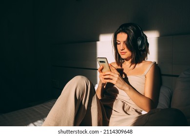 Young beautiful girl waking up in morning listening to her favorite music on headphones. Listening to music with your phone and wireless headphones on bed. - Shutterstock ID 2114309648