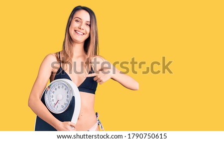 Young beautiful girl wearing bikini holding weighing machine smiling happy pointing with hand and finger 