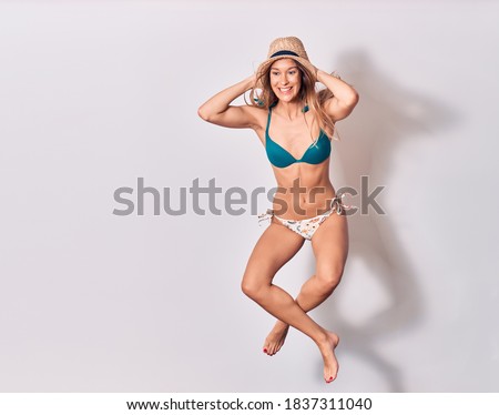 Young beautiful girl wearing bikini and summer hat smiling happy. Jumping with smile on face doing yoga position with legs crossed over isolated white background