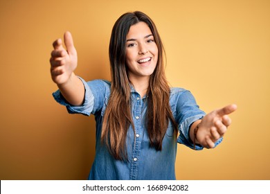 Young beautiful girl wearing casual denim shirt standing over isolated yellow background looking at the camera smiling with open arms for hug. Cheerful expression embracing happiness.
