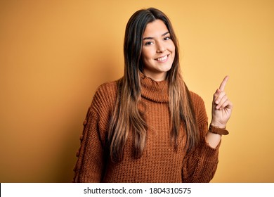 Young beautiful girl wearing casual sweater standing over isolated yellow background showing and pointing up with finger number one while smiling confident and happy.
