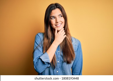 Young beautiful girl wearing casual denim shirt standing over isolated yellow background with hand on chin thinking about question, pensive expression. Smiling with thoughtful face. Doubt concept. - Shutterstock ID 1808193448