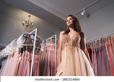 Young Beautiful Girl Wearing A Full-length Beige Yellow Chiffon Slit Prom Ball Gown Decorated With Golden Sparkles And Sequins. Fashion Model In Dress Hire Service With Many Dresses On Background.