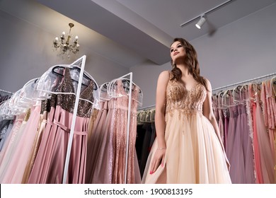 Young Beautiful Girl Wearing A Full-length Beige Yellow Chiffon Slit Prom Ball Gown Decorated With Golden Sparkles And Sequins. Fashion Model In Dress Hire Service With Many Dresses On Background.