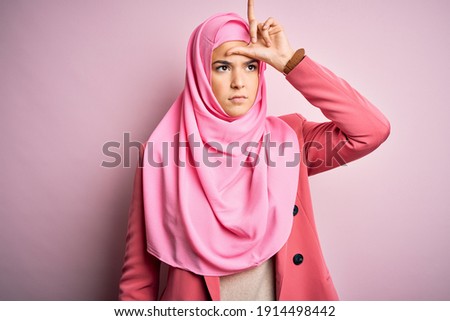 Young beautiful girl wearing muslim hijab standing over isolated pink background making fun of people with fingers on forehead doing loser gesture mocking and insulting.