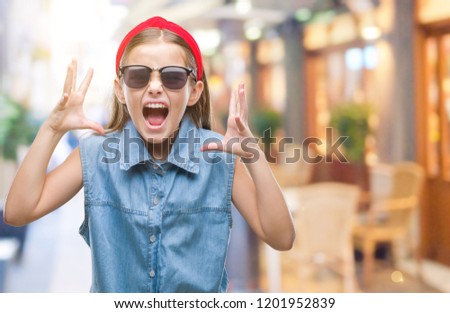 Young beautiful girl wearing sunglasses over isolated background crazy and mad shouting and yelling with aggressive expression and arms raised. Frustration concept.