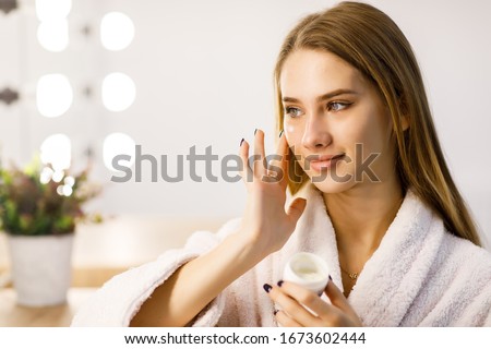 Young beautiful girl in a white robe cares for the skin using face cream. Beauty lifestyle.