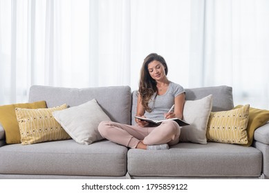 Young Beautiful Girl Writing A Love Letter To Her Boyfriend Or Writing In The Diary While She Is Smiling And Sitting On The Sofa In Her Room. Successful Female Having Business Ideas.