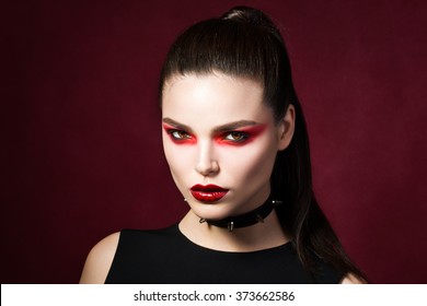Young beautiful gothic woman with white skin and red lips with bloody drops wearing black collar with spikes. Red smokey eyes. Vampire or halloween make-up.