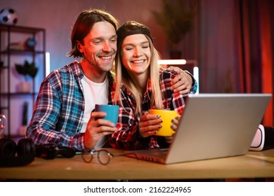 Young Beautiful Happy Couple Sitting At Wooden Table At Night Time In Living Room And Watching Movie And Drink Hot Drinks Tea Or Coffee In Mugs Using Laptop. Concept Of Evening Leisure And Technology.