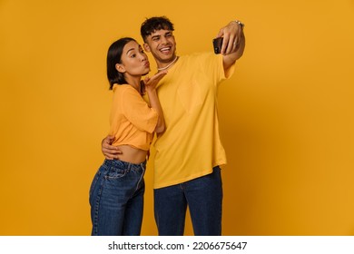 Young Beautiful Happy Couple Taking Selfie On Phone. Handsome Stylish Man Hugging His Asian Girlfriend With Puckered Lips