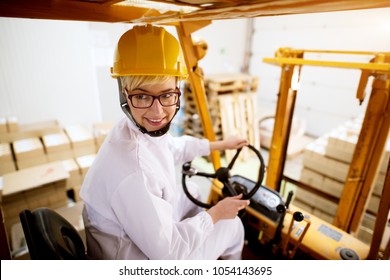 Young Beautiful Happy Female Worker Is Driving Fork Truck In Reverse While Carrying Pallet With A Stack Of Cardboard Boxes.