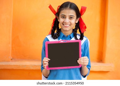 Young beautiful happy indian school girl holding slate against orange background, Smiling braided hair female teenager kid with black board. education concept. rural india. woman empowerment.