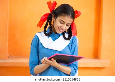 Young beautiful happy indian school girl write on slate against orange background, Smiling braided hair female teenager kid study with black board. education concept. rural india.
