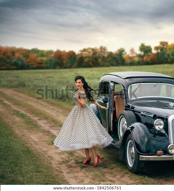 Young beautiful happy retro woman near old car.\
Attractive elegant lady in vintage white polka black dot retro\
dress. Pin-up hairstyle. Cheerful girl standing on road. Autumn\
nature, yellow trees