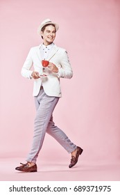 Young Beautiful Happy Smiling Modern Male Model Wearing White Jacket, White Shirt, Grey Pants And White Hat Stepping Through Studio With Light Pink Background, Holding Red Coffee Cup And Looking Away