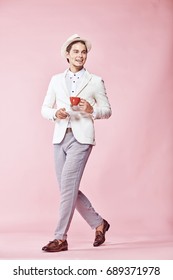 Young Beautiful Happy Smiling Modern Man Wearing White Jacket, White Shirt, Grey Pants And White Hat Stepping Through Studio With Light Pink Background, Holding Red Coffee Cup And Looking Away