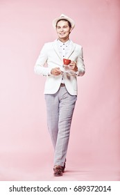 Young Beautiful Happy Smiling Modern Man Wearing White Jacket, White Shirt, Grey Pants And White Hat Stepping Through Studio With Light Pink Background, Holding Red Coffee Cup And Looking Away