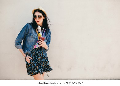young beautiful happy stylish hipster girl, cocktail, smoozy drink, denim jacket, smiling, fashion, teen, cool accessories, purse, hat, sunglasses, amazed, vintage style, wall background, hair, wind