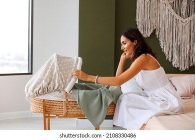 Young beautiful happy woman mother looking at her little newborn baby sleeping in bed bassinet at home, smiling mom communicating with child lying in crib. Motherhood concept