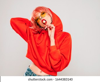 Young beautiful hipster bad girl in trendy red summer red hoodie and earring in her nose.Sexy carefree woman posing in studio on gray background in wig.Hot model licking round sugar candy - Shutterstock ID 1644383140
