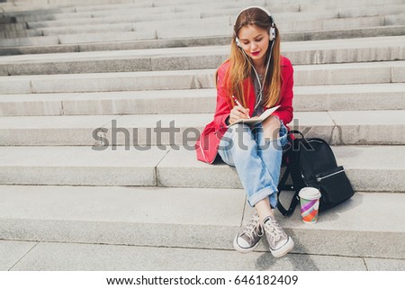 young beautiful hipster blond woman, pink coat, jeans, sitting on stairs,  spring summer fashion, urban style, backpack, smiling, happy, positive, listen to music, headphones, travel diary, writing 
