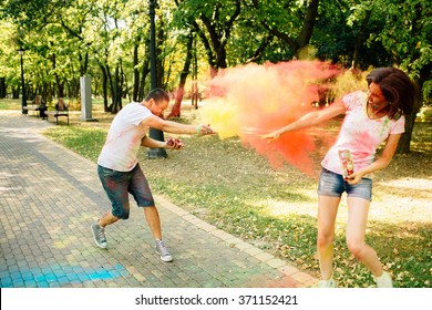 Young And Beautiful, Hipster Couple Funny Playing In The Park On Holi Color Festival Throw Each Other With Colour Paint Powder. Dressed In White T-shirts And Denim Shorts.