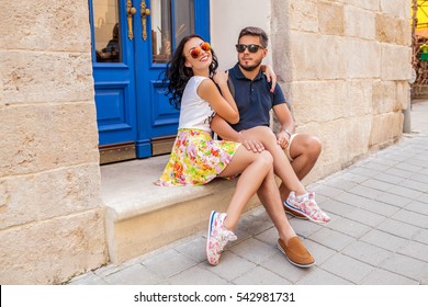 young beautiful hipster couple in love sitting on old city street, summer Europe vacation, travel, fun, happy, smiling, sunglasses, trendy outfit, romance, date, embracing, legs, sneakers