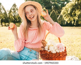 Young beautiful hipster woman in trendy summer jeans, pink T-shirt and hat. Woman making picnic outside. Positive model sitting on plaid on grass, eating fruits and cheese, holding lemons in hands 