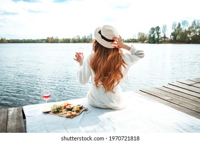 Young beautiful hipster woman in trendy summer sundress and hat. Carefree woman making picnic outside. Positive model sitting on blanket, eating fruits, drinking rose wine