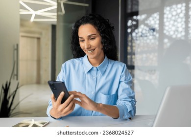 Young beautiful hispanic businesswoman at workplace inside office, woman smiling contentedly looking at phone screen, using smartphone app, typing messages, online social networks.