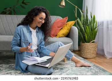 Young beautiful Hispanic female student studying remotely online at home, taking lecture notes sitting on floor in living room with green wall, woman taking courses studying online - Powered by Shutterstock