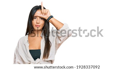 Young beautiful hispanic girl wearing casual clothes making fun of people with fingers on forehead doing loser gesture mocking and insulting. 