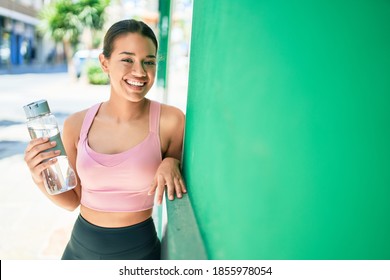 Young beautiful hispanic sporty woman wearing fitness outfit smiling happy and natural leaning on a green chroma wall at the town drinking fresh water from the bottle