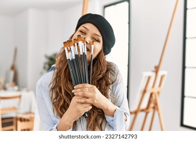 Young beautiful hispanic woman artist covering mouth with paintbrushes at art studio - Powered by Shutterstock