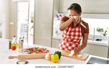Young beautiful hispanic woman cutting onion crying at the kitchen