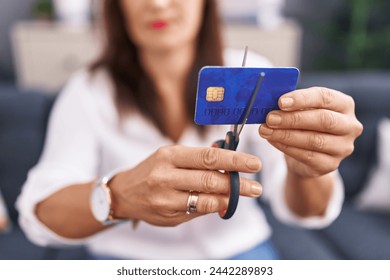 Young beautiful hispanic woman cutting credit card sitting on sofa at home - Powered by Shutterstock