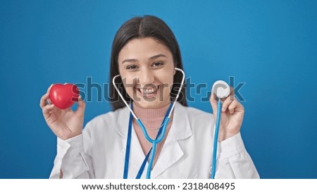 Young beautiful hispanic woman doctor smiling confident holding stethoscope and heart over isolated blue background