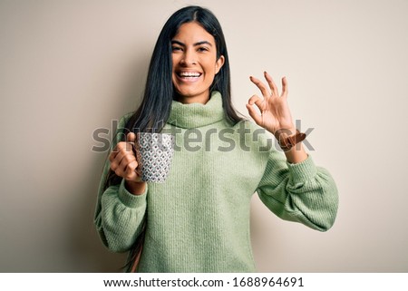 Young beautiful hispanic woman drinking a cup of hot coffee over isolated background doing ok sign with fingers, excellent symbol
