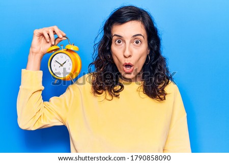 Young beautiful hispanic woman holding alarm clock scared and amazed with open mouth for surprise, disbelief face 