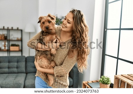 Young beautiful hispanic woman hugging dog standing at home