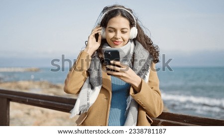 Young beautiful hispanic woman listening to music with smartphone at seaside