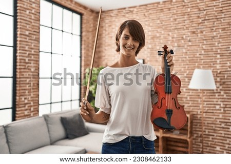 Young beautiful hispanic woman musician smiling confident holding violin at home