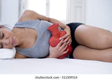 Young Beautiful Hispanic Woman In Painful Expression Holding Hot Water Bottle Against Belly Suffering Menstrual Period Pain Lying Sad On Home Bed Having Tummy Cramp In Female Health Concept